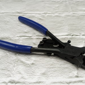 Puede incluir: Una crimpadora negra con mangos azules, diseñada para trabajos eléctricos. La herramienta está abierta, mostrando sus mordazas y el mecanismo de crimpado. Los mangos azules contrastan con el cuerpo negro.