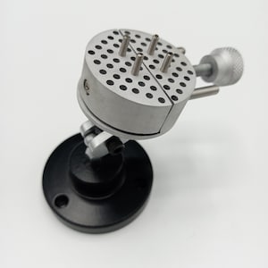 May include: A silver metal watchmaker's vice with a black base. The vise has a rotating head with multiple holes and pins for holding small objects.
