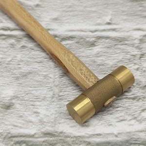 May include: A brass hammer with a wooden handle. The hammer has a textured head and a smooth handle.