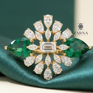 May include: A gold ring featuring a central emerald-cut diamond surrounded by pear-shaped diamonds and two large, pear-shaped green gemstones. The ring has a floral design and is set against a green background. The ZARNA JEWELS logo is visible.