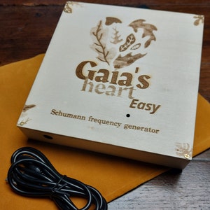 Könnte beinhalten: Eine Holzkiste mit einem herzförmigen Design und dem Text "Gaia's heart Easy Schumann frequency generator" auf der Oberseite. Die Kiste ist hellbraun und hat ein schwarzes Stromkabel.