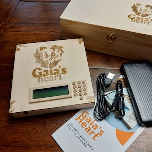 Può includere: Una scatola di legno con un logo Gaia's Heart e un piccolo dispositivo elettronico con uno schermo e pulsanti. Il dispositivo è collegato a un adattatore di corrente e a un cavo. La scatola è aperta e il dispositivo è all'interno. La scatola è su un tavolo di legno.