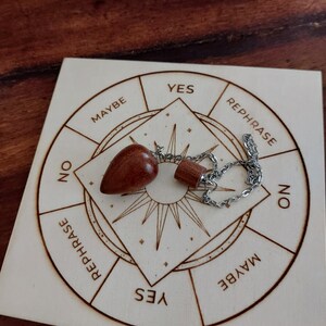 Può includere: Un pendolo di legno con una catena, appoggiato su un pannello di legno con un grafico circolare. Il grafico ha le parole "Sì", "No", "Forse" e "Riformulare" scritte attorno al bordo.