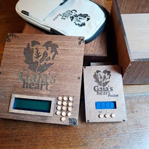 Könnte beinhalten: Ein elektronisches Gerät aus dunklem Holz mit digitalem Bildschirm, Ziffernblock und der lasergravierten Aufschrift Gaia's heart. Daneben liegt ein kleineres Holzgerät mit der Aufschrift Gaia's heart Pocket, blauem Display und beiger Stofftasche.