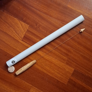 Op de afbeelding: Een witte kartonnen buis met een houten deuvel en een klein houten pijlvormig object bevestigd aan een touwtje. Een houten schijf en een houten handvat zijn ook aanwezig. De items liggen op een houten oppervlak.