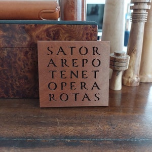 Può includere: Un sottobicchiere quadrato in legno con la frase latina "SATOR AREPO TENET OPERA ROTAS" incisa su di esso.