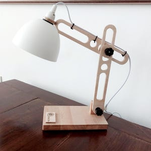 Könnte beinhalten: Eine Schreibtischlampe aus Holz mit einem weißen Schirm und einem schwarzen Knopf. Die Lampe steht auf einem Holzsockel mit der eingravierten Aufschrift "DESIGN".