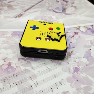 May include: A yellow and black handheld game console with a Pikachu character on the front. The console has a black screen and a yellow button with a plus sign on it. The console is labeled "Pokemon".