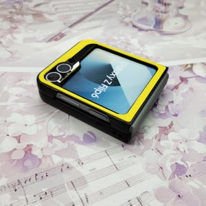May include: A yellow and black phone case with a black screen that says "Galaxy Z Flip" in white text. The case has a camera lens on the back.