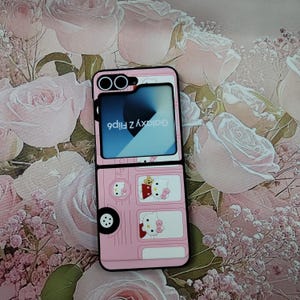 May include: A pink phone case with a Hello Kitty design. The case is designed to look like a bus with Hello Kitty characters on the windows. The phone case is for a Samsung Galaxy Z Flip 4.