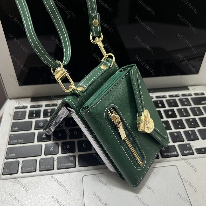 Puede incluir: Una cartera de cuero verde con una cremallera dorada y un dije dorado en forma de coraz&oacute;n. La cartera tiene una correa que se puede usar para llevarla como bolso de bandolera.