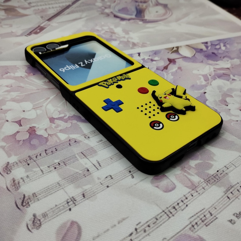 May include: A yellow phone case with a Pokemon Game Boy design. The case has a black border and a Pikachu character on the side. The screen is visible and shows the text "flip Z AxorPro".