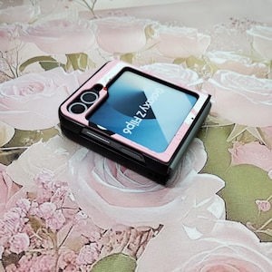 May include: A pink and black Galaxy Z Flip phone case with a clear back panel showing the phone's screen. The phone's screen displays the text "Galaxy Z Flip".