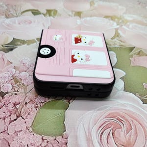May include: Pink Hello Kitty phone case with a black and white bus design. The case has a black trim and a Hello Kitty face on the front.