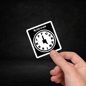 May include: A black and white sticker featuring the text "My clock at work" above a clock face. The clock face has the words "IT FUCK THIS" in place of numbers. The sticker is held by a hand.