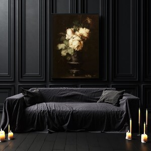 May include: A black sofa with a dark gray throw blanket and two pillows. A framed painting of white roses in a vase hangs on the wall behind the sofa. There are candles on the floor in front of the sofa.