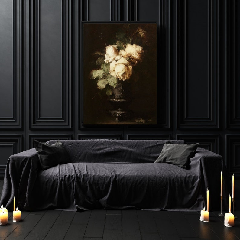 May include: A black sofa with a dark gray throw blanket and two pillows. A framed painting of white roses in a vase hangs on the wall behind the sofa. There are candles on the floor in front of the sofa.