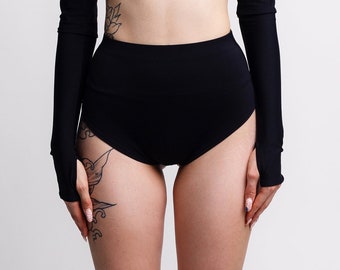 May include: A black two-piece outfit consisting of a high-neck crop top, high-waisted shorts, and fingerless sleeves. The model is wearing black platform boots with multiple buckles. The outfit is a sleek, modern style.