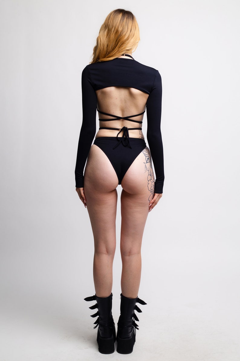 May include: Black long-sleeved crop top with a criss-cross back design and matching high-waisted bikini bottoms. The model is wearing black platform boots.