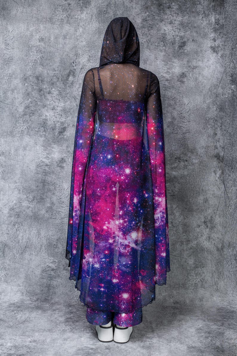 May include: A sheer black hooded top with a galaxy print. The top is long-sleeved and has a flowing, cape-like design. The print features a dark blue background with pink, purple, and white stars.