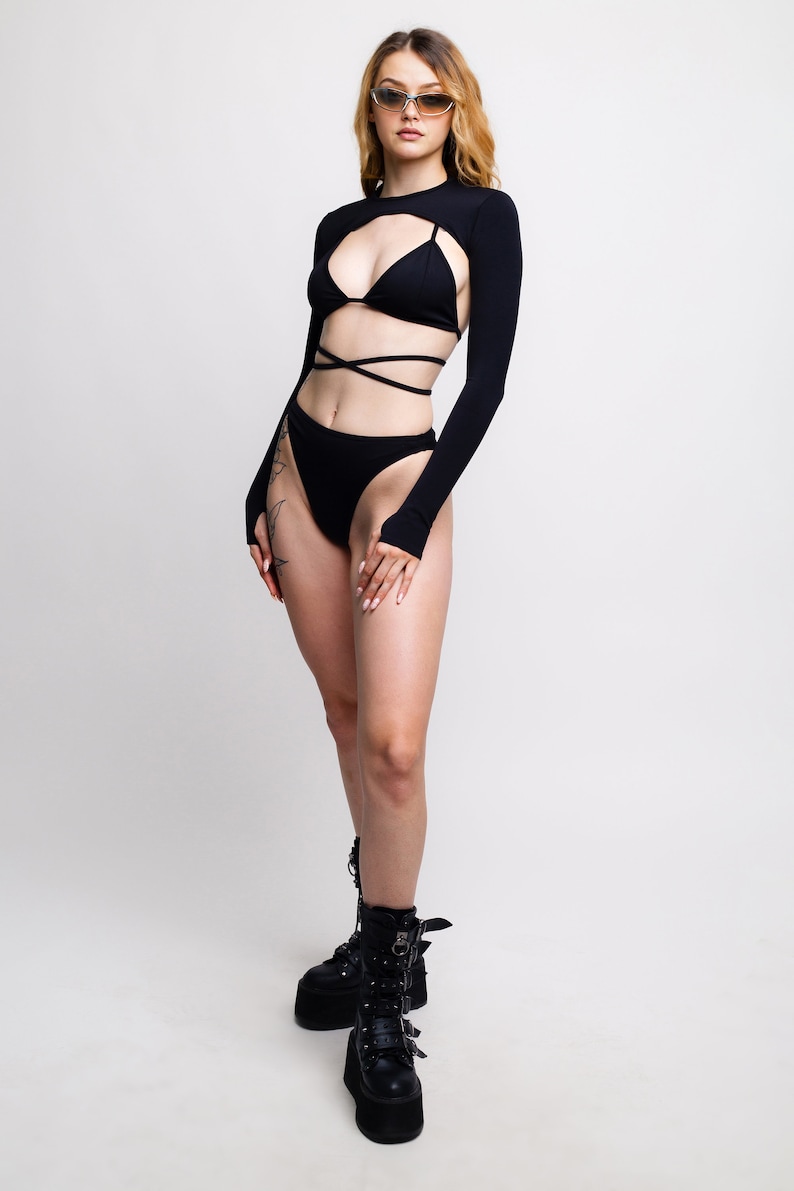 May include: A woman wearing a black long-sleeved crop top with a cut-out design and a black bikini bottom. She is wearing black platform boots with buckles.