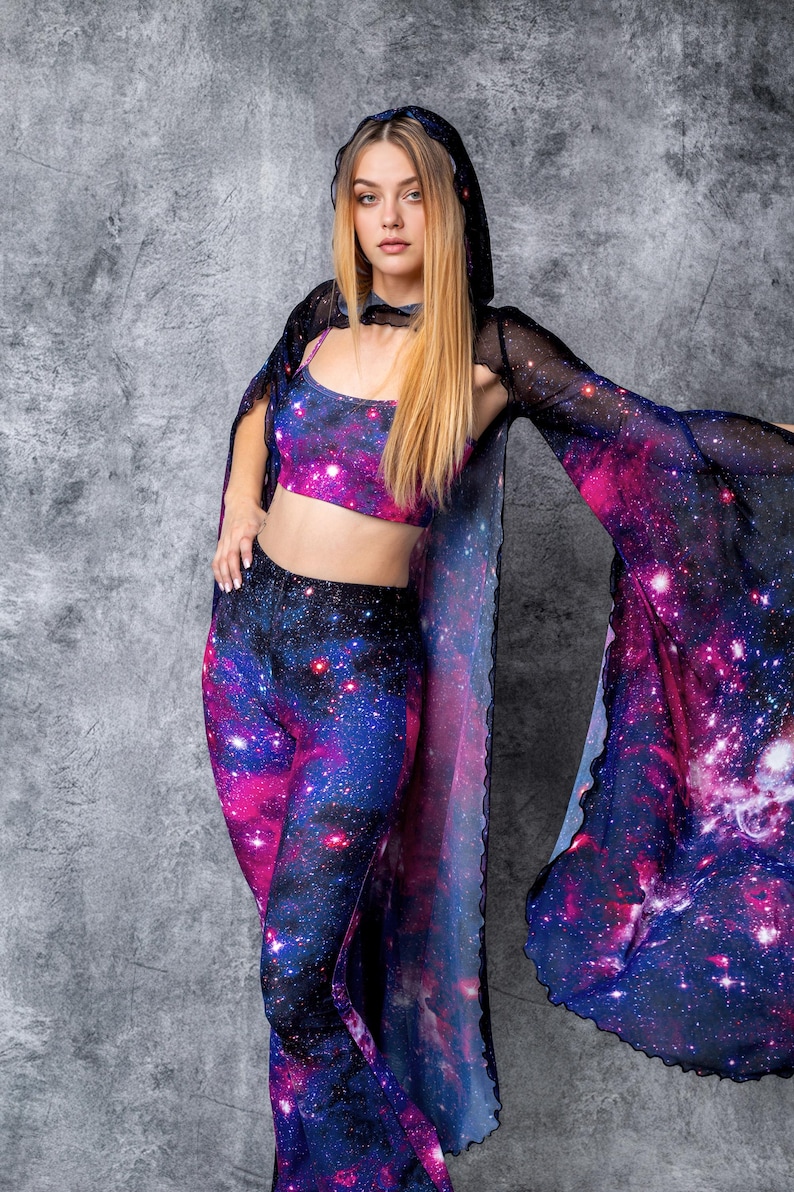 May include: A woman wearing a galaxy print hooded cape, crop top, and bell bottom pants. The outfit is made of a sheer, stretchy fabric with a galaxy print.