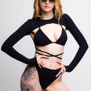 May include: A woman wearing a black crop top with long sleeves and a black bikini bottom. She has a large floral tattoo on her left thigh. She is wearing sunglasses and has her hair down.