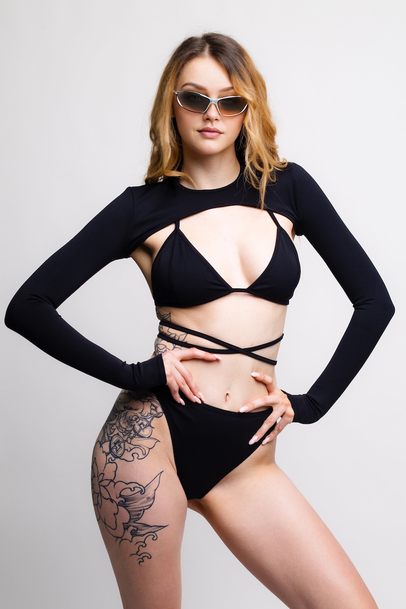 May include: A woman wearing a black crop top with long sleeves and a black bikini bottom. She has a large floral tattoo on her left thigh. She is wearing sunglasses and has her hair down.