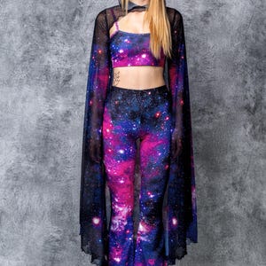 May include: A woman wearing a black mesh cape with galaxy print bell bottom pants and a galaxy print crop top. She is wearing white platform shoes.