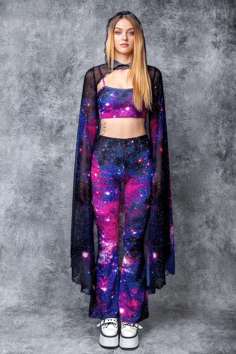 May include: A woman wearing a black mesh cape with galaxy print bell bottom pants and a galaxy print crop top. She is wearing white platform shoes.