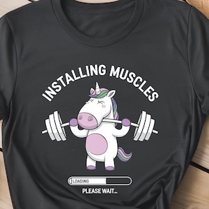 May include: A black t-shirt with a white graphic of a unicorn lifting a barbell. The text "Installing Muscles" is above the unicorn and "Loading... Please wait..." is below the unicorn.