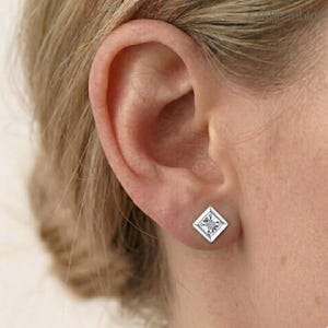 Könnte beinhalten: Ein silberfarbener Ohrstecker mit quadratischem Rahmen und einem runden, klaren Stein in der Mitte. Der Ohrstecker befindet sich an einem Ohrläppchen und zeigt ein geometrisches Design mit einer polierten, metallischen Oberfläche und einem facettierten Stein.