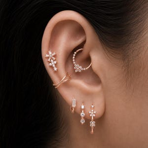 Könnte beinhalten: Eine Kollektion von Ohrringen aus Roségold mit klaren Steinen. Das Set enthält einen blumenförmigen Stecker, einen Kreuzstecker, eine schlichte gekreuzte Manschette, einen Daith-Ring mit Blumenmotiv und drei kleine Creolen mit Steinen.