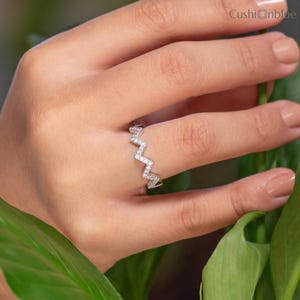 Könnte beinhalten: Ein silberfarbener Ring mit einem zickzack- oder chevronförmigen Band, das mit kleinen, klaren, funkelnden Steinen besetzt ist. Der Ring hat eine polierte metallische Oberfläche und ein geometrisches Design. Der Text Cushionblue ist oben rechts sichtbar.