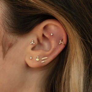 Könnte beinhalten: Nahaufnahme eines Ohres mit mehreren goldenen Ohrringen. Die Ohrringe umfassen einen kleinen Diamanten-Stecker, einen kleinen goldenen Kreis, einen kleinen goldenen Stab, einen kleinen goldenen Kreis und einen dreizackigen Ohrring mit drei kleinen Diamanten.