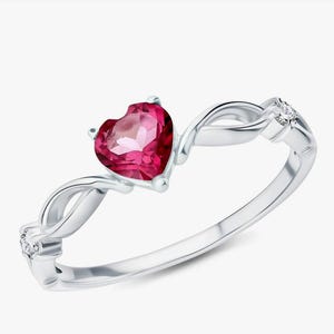 Könnte beinhalten: Ein silberner Ring mit einem herzförmigen, leuchtend roten Edelstein. Der Ring hat ein verdrehtes Banddesign mit kleinen Akzentsteinen auf beiden Seiten des herzförmigen Steins. Der Ring ist vor einem weißen Hintergrund platziert.