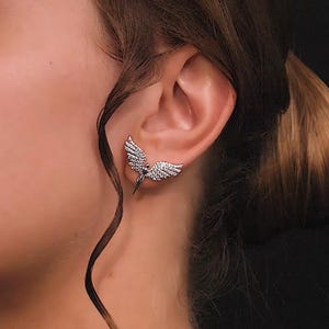 May include: Silver angel wing earrings with clear cubic zirconia stones. The wings are slightly angled and the earrings are worn in the earlobe.
