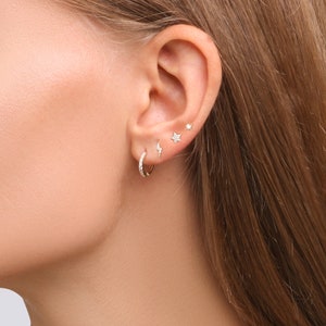 May include: A close-up of a person's ear with multiple earrings. The earrings include a small hoop, a lightning bolt stud, and two star studs.