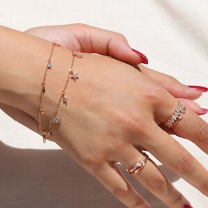 Könnte beinhalten: Ein Set aus zarten goldfarbenen Armbändern mit baumelnden Anhängern, darunter ein Halbmond, ein Stern und ein Blitz. Die Armbänder sind übereinander auf einem Handgelenk gelegt und erzeugen einen stilvollen und verspielten Look.