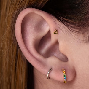 May include: Close-up of an ear with three earrings. A small gold triangle stud is in the upper cartilage. A silver hoop earring is in the lobe, and a gold hoop with rainbow-colored gemstones is in the lower lobe.
