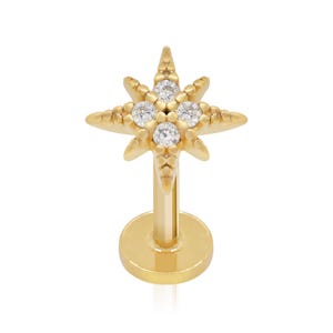 Könnte beinhalten: Ein goldfarbener Ohrstecker mit flacher Rückseite und einem sternförmigen Aufsatz. Das Sterndesign ist mit vier kleinen, klaren, rund geschliffenen Steinen in einer zentralen Anordnung verziert, umgeben von texturierten, spitzen Strahlen.