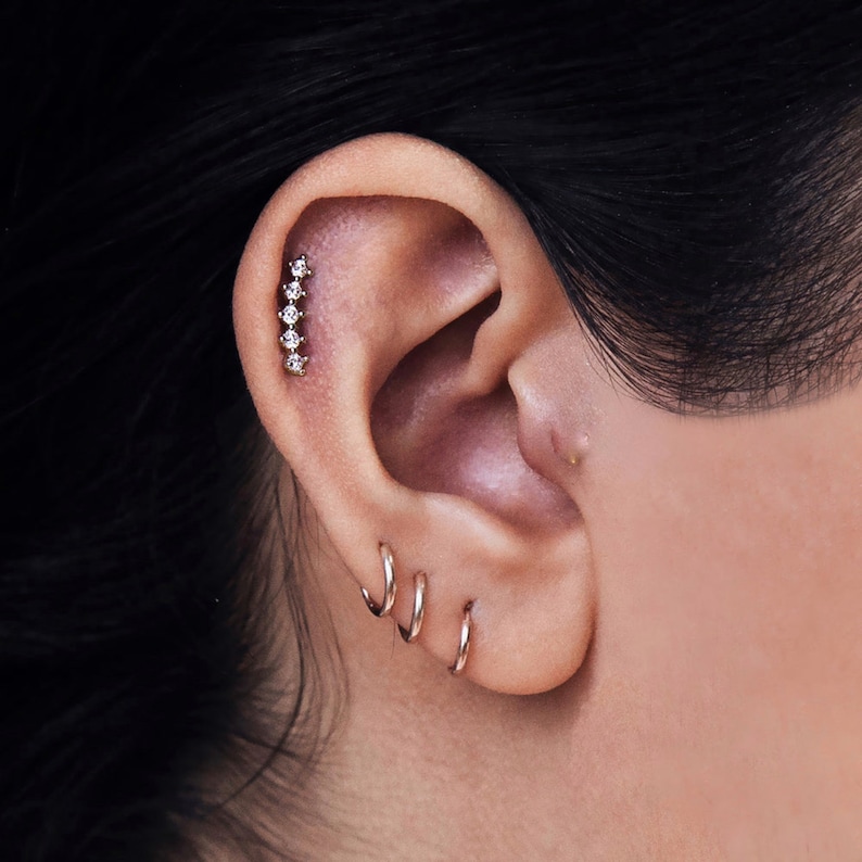 May include: A close-up of a person's ear with multiple piercings. The ear is pierced with a row of small diamonds in the helix and three gold hoops in the lobe.