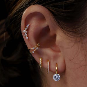 Könnte beinhalten: Ein goldenes Ohr-Set bestehend aus einem geschwungenen Climber-Ohrring mit marquise-geschliffenen Steinen, einer doppelten Ohrklemme mit kleinen runden Steinen, einem Perlen-Creolen, einem schmalen Pavé-Creolen und einem Creolen mit rundem Anhänger.