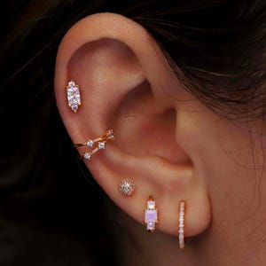 Könnte beinhalten: Ein goldfarbenes Ohr-Set bestehend aus einem Stecker mit rechteckigem klaren Stein, einem doppelten Creolen-Reif mit kleinen klaren Steinen, einem strukturierten Goldkugel-Stecker, einer kleinen Creole mit quadratischem Opalstein und zwei klaren Steinen.