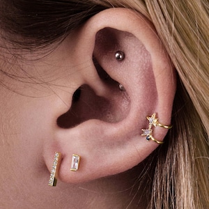 Könnte beinhalten: Nahaufnahme eines Ohrs mit mehreren Ohrringen. Das Ohr hat ein Helix-Piercing mit einem kleinen silbernen Stecker, ein Ohrläppchen-Piercing mit einem dünnen goldenen Stab, der mit klaren Steinen besetzt ist, und ein Conch-Piercing mit einem goldenen Ohrring in Form eines Sterns mit klaren und blauen Steinen.