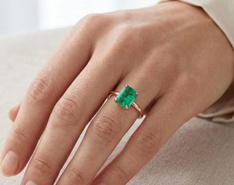 Op de afbeelding: Een ring met een rechthoekige smaragdgeslepen groene edelsteen in een gepolijste gouden band. De ring zit om een vinger en toont de levendige groene steen die door vier pootjes wordt vastgehouden. De tekst Cushionblue is in de hoek zichtbaar.