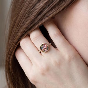 Könnte beinhalten: Ein goldener Ring mit einer kreisförmigen Fassung, die mit einer Ansammlung kleiner, mehrfarbiger Edelsteine in Rosa-, Blau- und Grüntönen gefüllt ist. Das Band ist dünn und metallisch, und der Text Cushionblue ist oben rechts zu sehen.