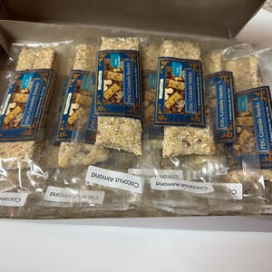 Może przedstawiać: Pudełko z indywidualnie zapakowanymi batonami granola. Każdy baton ma niebiesko-złotą etykietę z napisem "PDG Granola Snacks". Batony są oznaczone jako "Coconut Almond".
