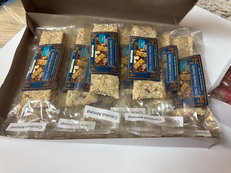 Może przedstawiać: Pudełko z indywidualnie zapakowanymi batonami granola. Każdy baton ma niebiesko-złotą etykietę z napisem "PDG Granola Snacks". Batony są oznaczone jako "Coconut Almond".