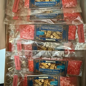 Może przedstawiać: Pudełko wypełnione pojedynczo zapakowanymi przekąskami PDG Granola, oznaczone jako "Fruit Almond". Każdy baton ma żywy czerwony kolor i jest zapakowany w przezroczysty plastik. Etykiety mają niebiesko-złoty wz&oacute;r z tekstem.
